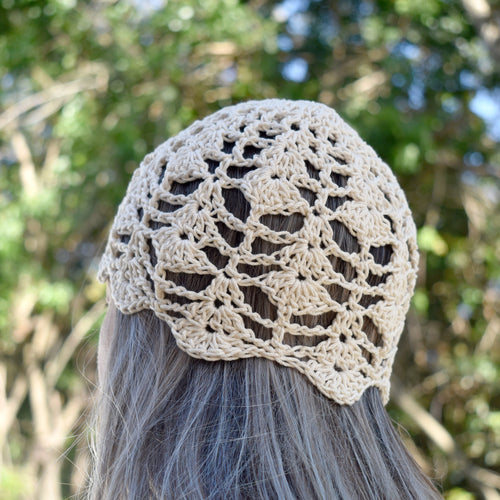 A person wearing a beige crochet skull cap against blurred green foliage in the background.