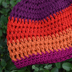 Close-up of a colorful knitted hat with purple, orange, and red stripes on a green leaf background.