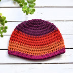 Colorful red, purple, and orange crocheted beanie on a white wooden surface with green plants.