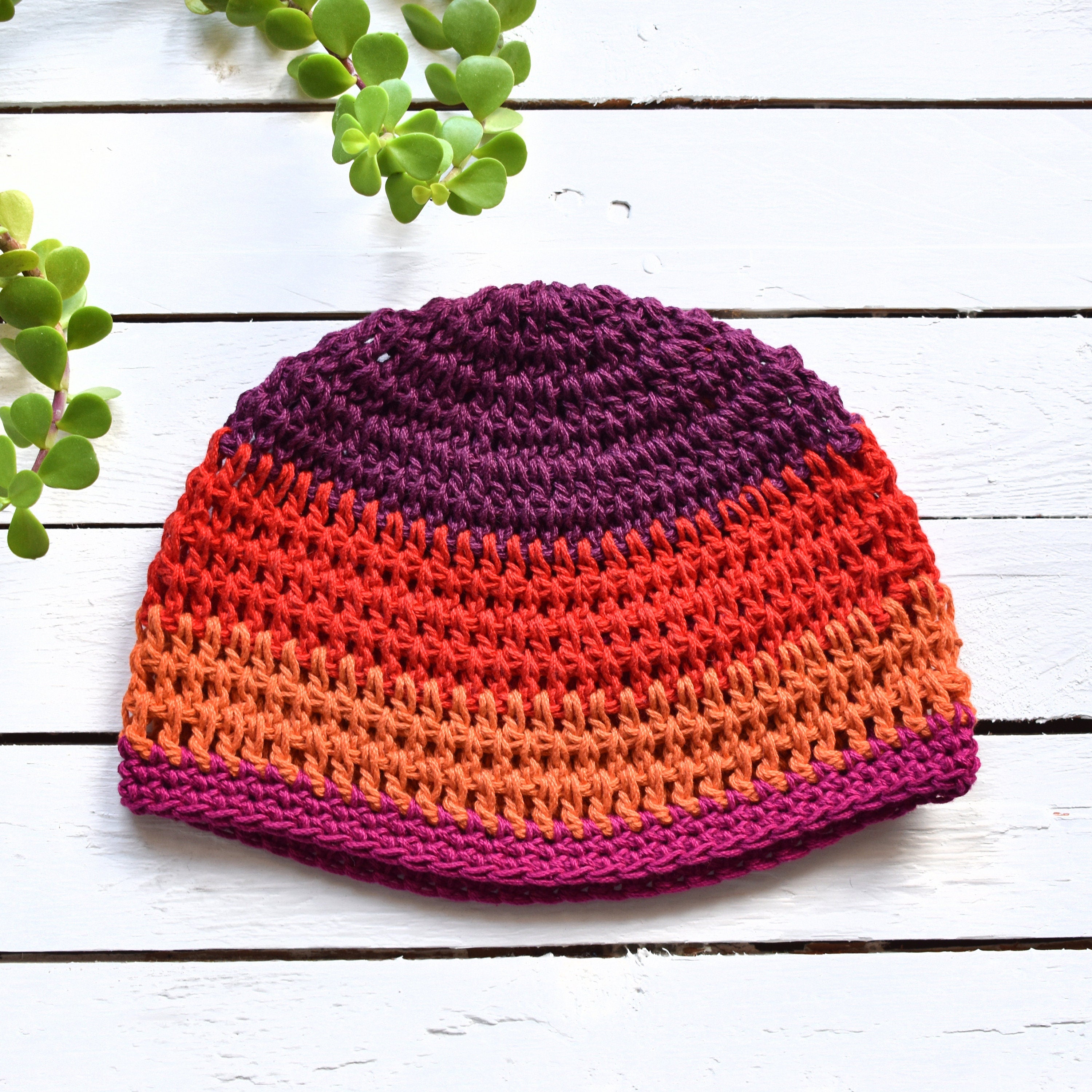 Colorful red, purple, and orange crocheted beanie on a white wooden surface with green plants.