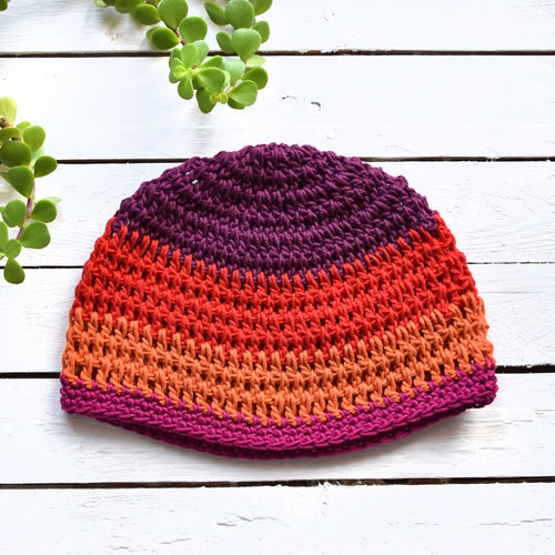 Colorful red, purple, and orange crocheted beanie on a white wooden surface with green plants.