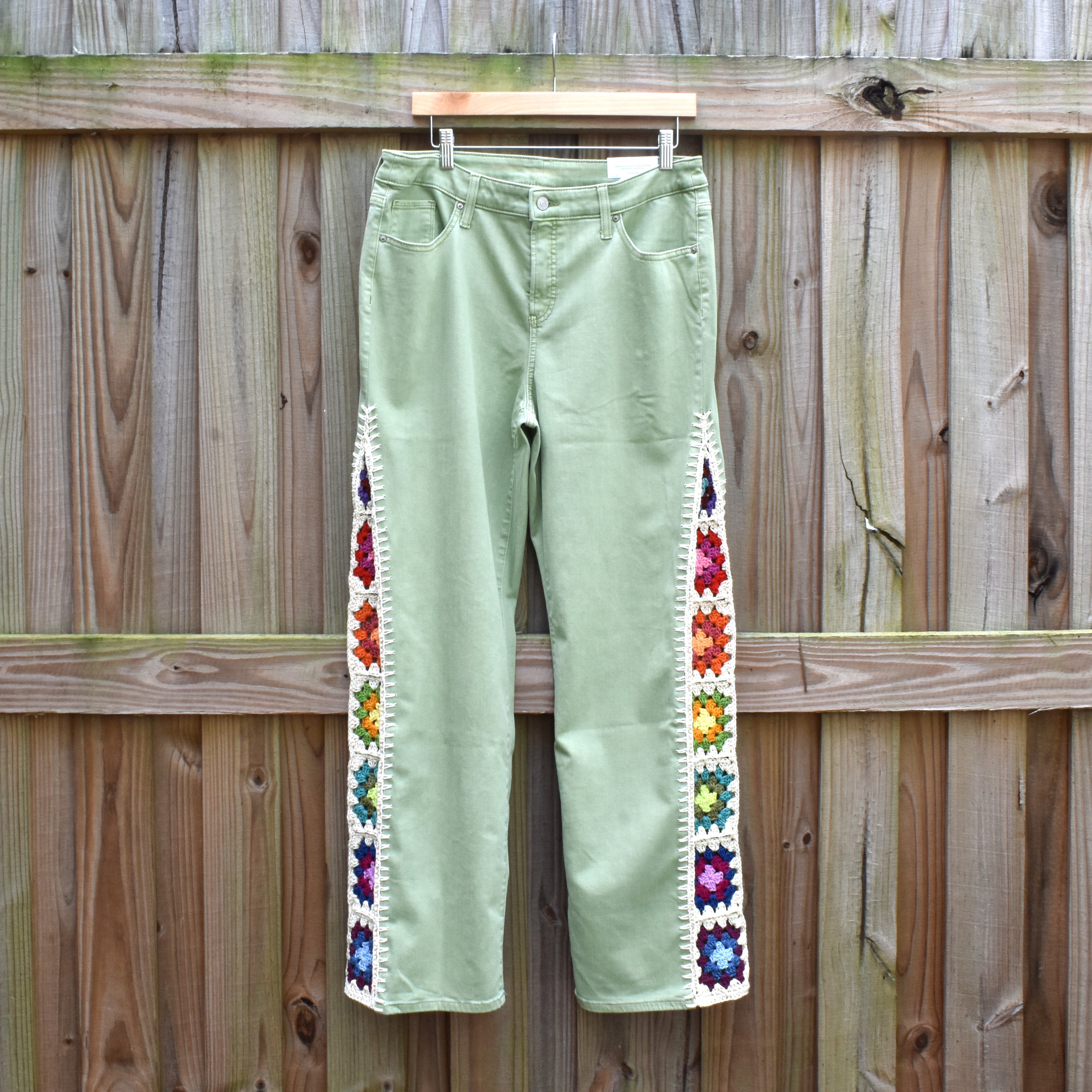 Light green Chico pants with colorful granny square inserts on the outside of each leg hanging on a wooden fence.