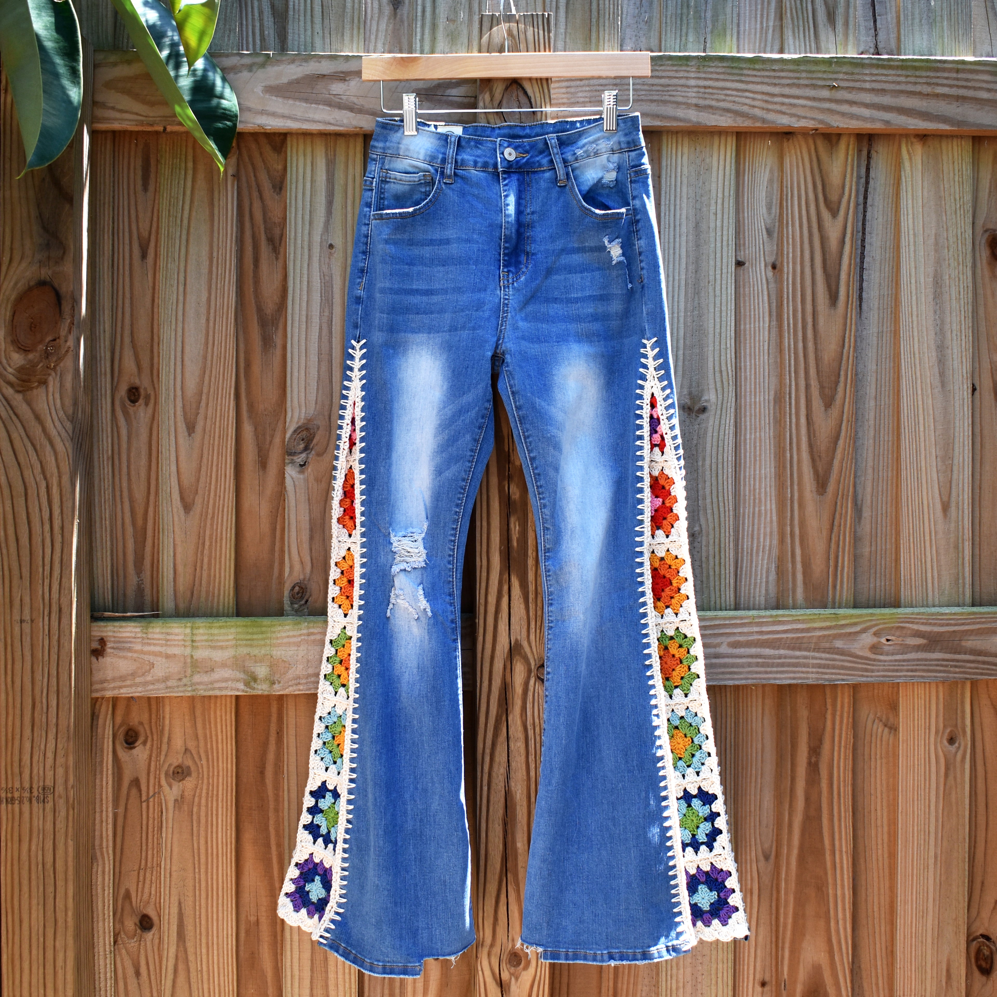 Blue flare jeans with colorful crochet granny square inserts hanging on a wooden fence.