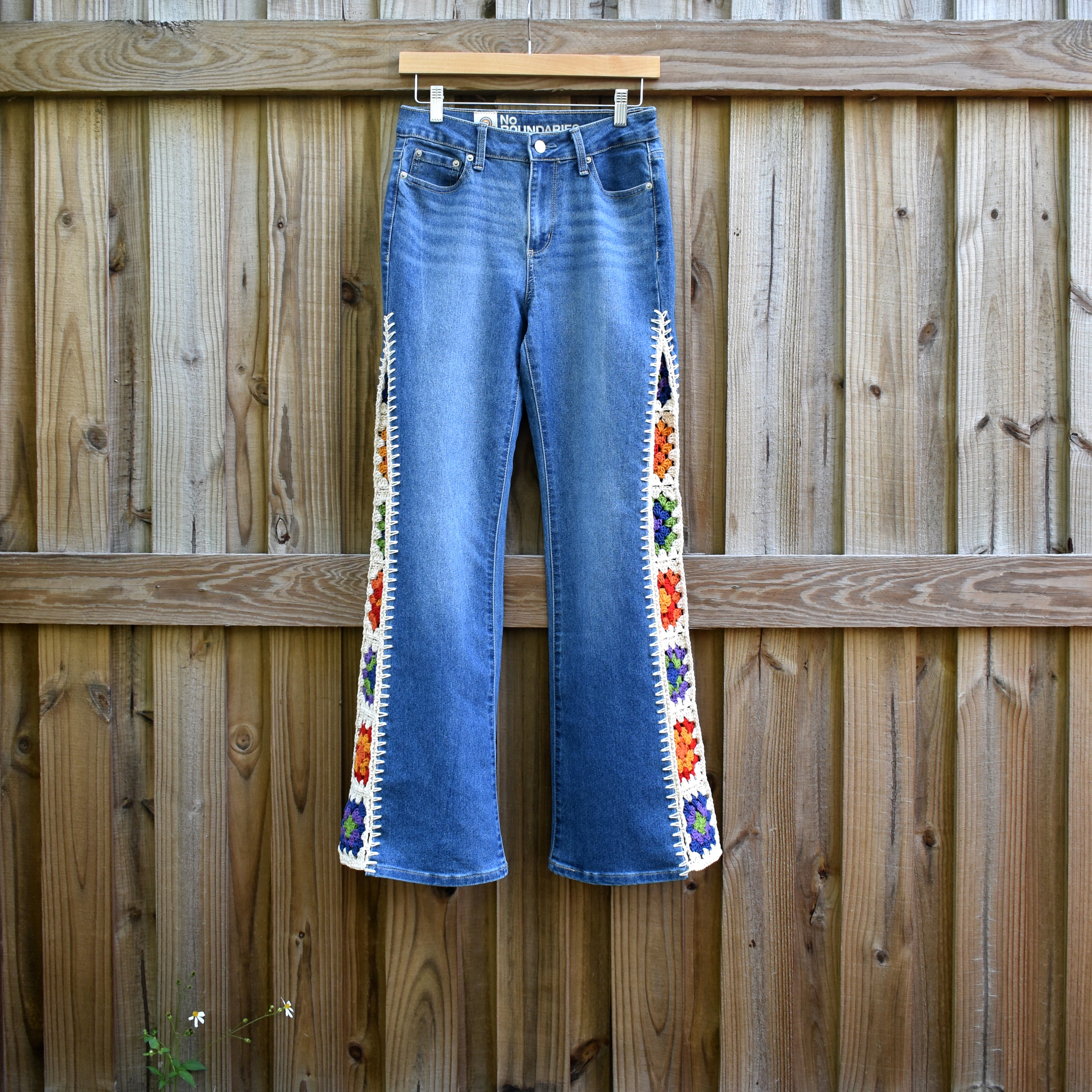 A pair of No Boundaries bootcut blue jeans with colorful crochet granny square inserts hanging on a wooden fence.