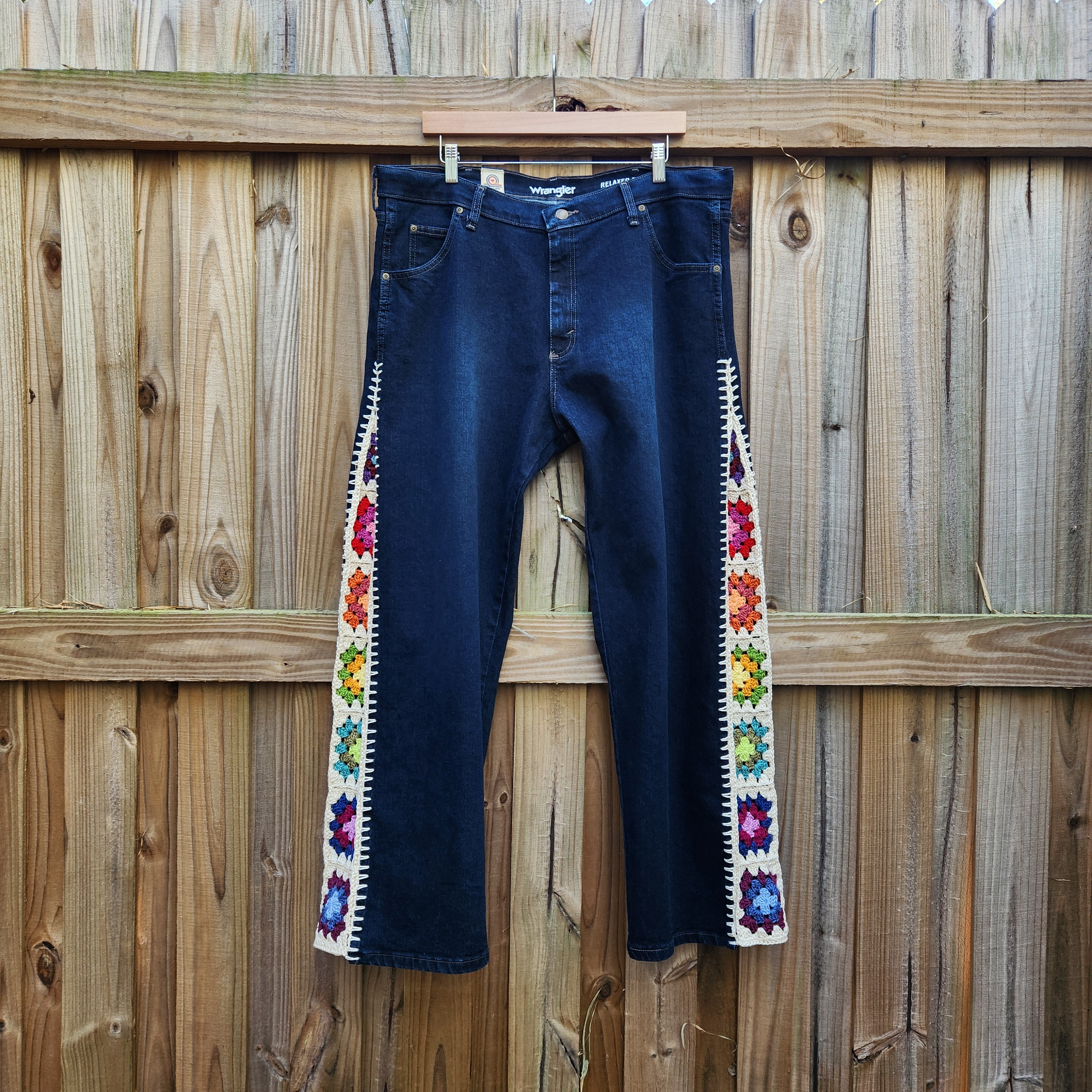 A pair of Wrangler blue jeans with colorful crochet granny square inserts on the outside of each leg hanging on a wooden fence.