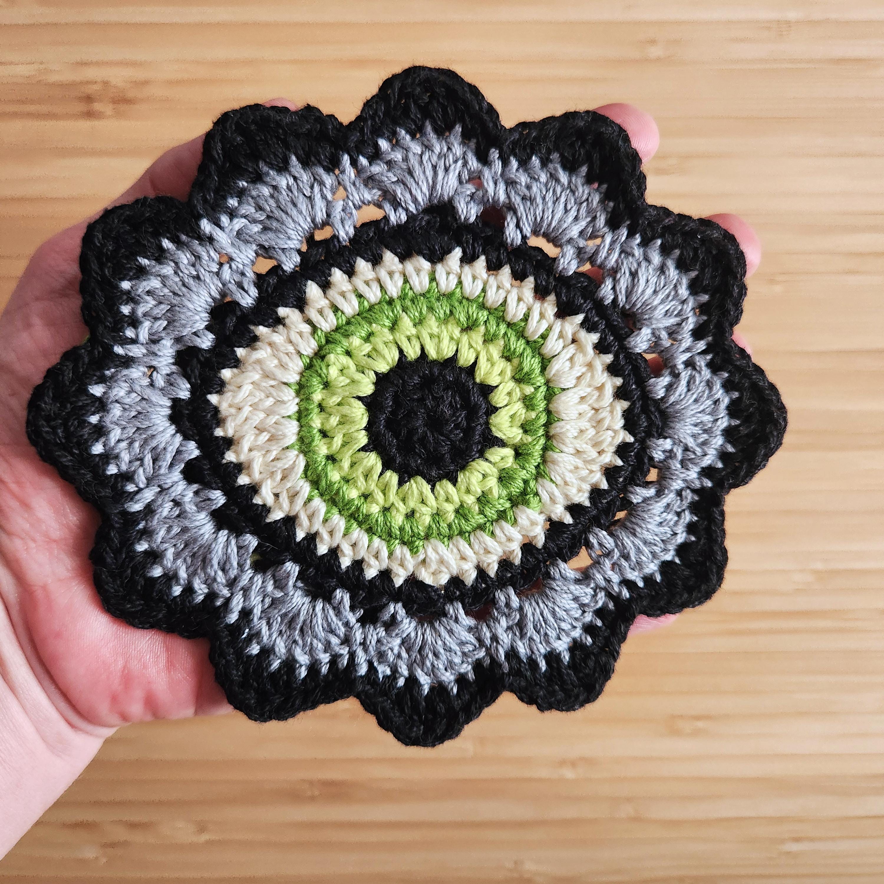 A hand holding a gray and black crochet flower with a lime green eye in the center against a wooden background.
