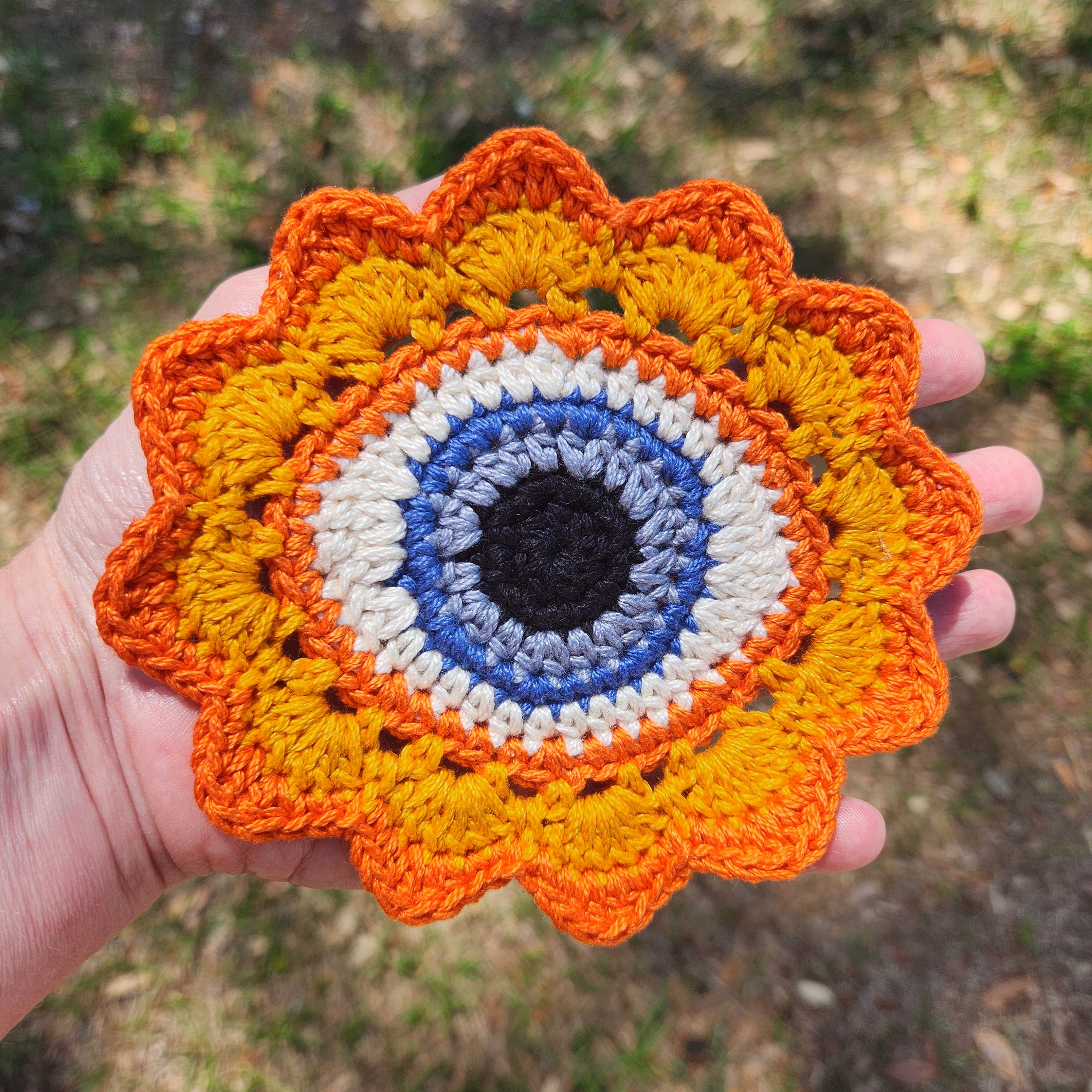 Hand holding a bright yellow crochet flower patch with an orange trim and a blue eye in the middle.