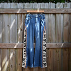 Blue denim flare jeans with black and white geometric patterned side panels, hanging on a wooden hanger against a wooden fence.