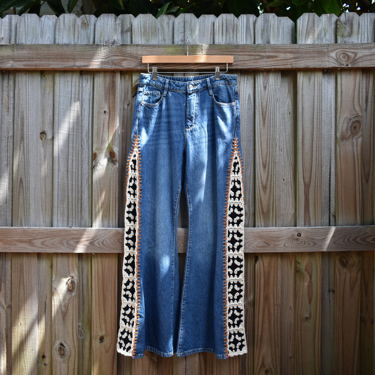 Blue denim flare jeans with black and white geometric patterned side panels, hanging on a wooden hanger against a wooden fence.