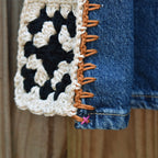 Close-up of a crocheted granny square patch in cream, black, and white, attached to a blue denim garment with orange-brown whipstitching, against a wooden background.