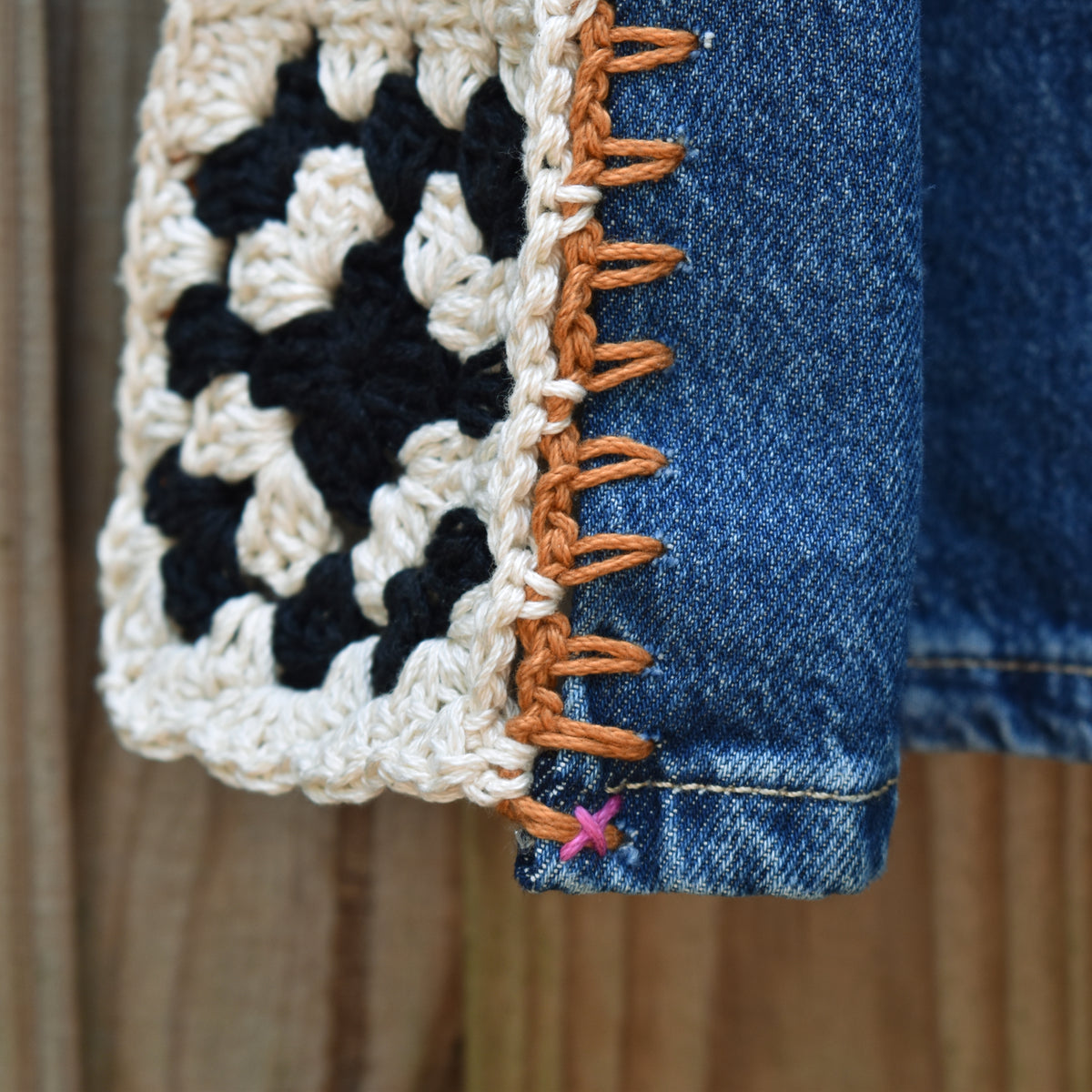 Close-up of a crocheted granny square patch in cream, black, and white, attached to a blue denim garment with orange-brown whipstitching, against a wooden background.