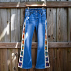 A pair of blue denim jeans hanging on a wooden fence with crochet rainbow granny square inserts running down the outside of each leg.
