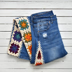 A pair of blue denim jeans with colorful hand-crocheted granny square and triangle patches attached to the sides, laid flat on a white wooden plank background.