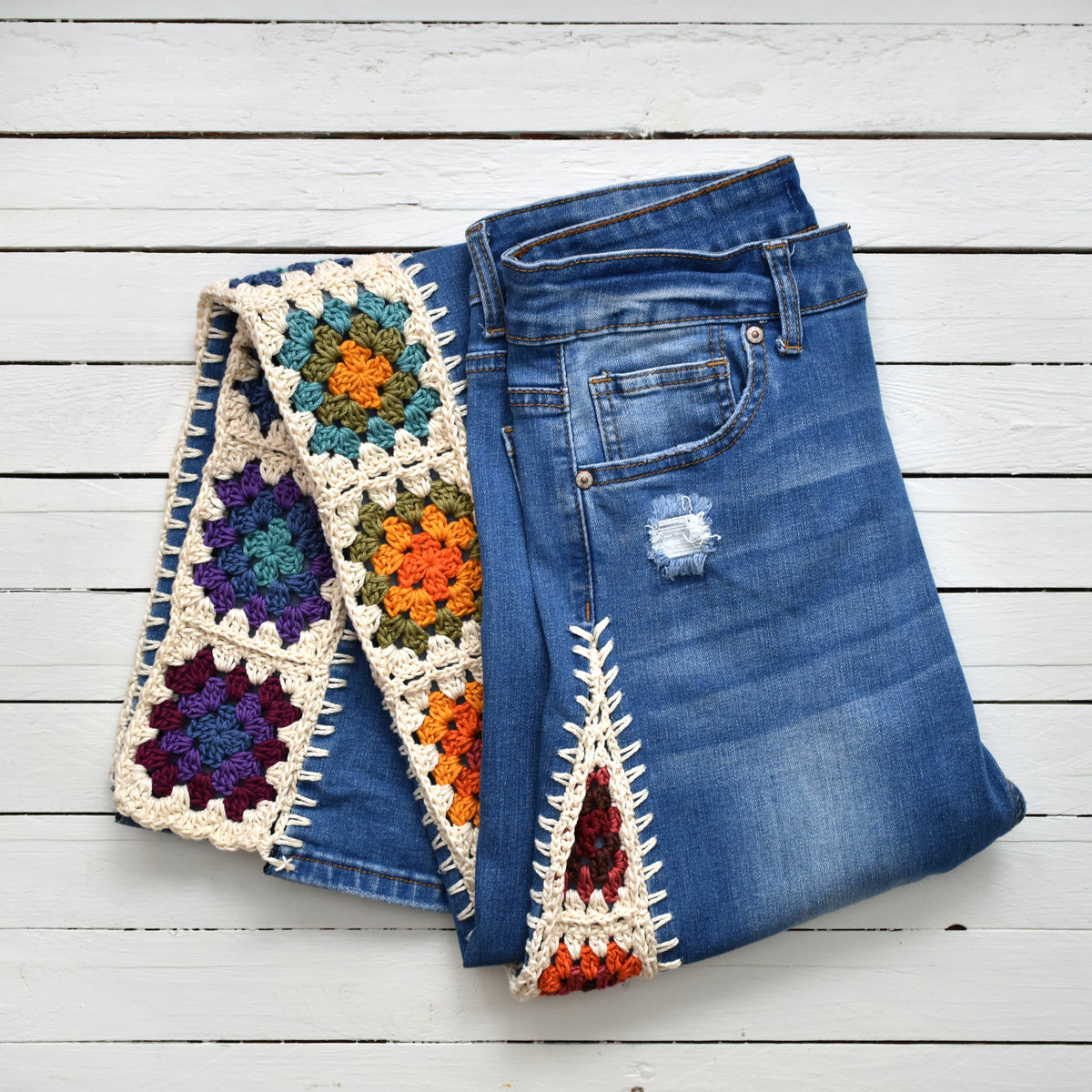 A pair of blue denim jeans with colorful hand-crocheted granny square and triangle patches attached to the sides, laid flat on a white wooden plank background.