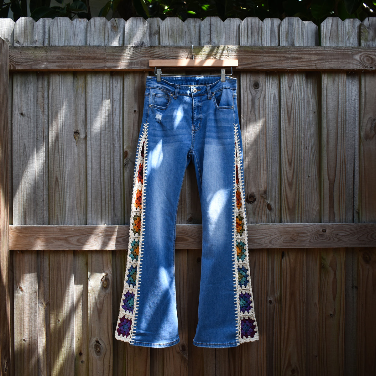A pair of blue denim jeans hanging on a wooden fence with crochet rainbow granny square inserts running down the outside of each leg.