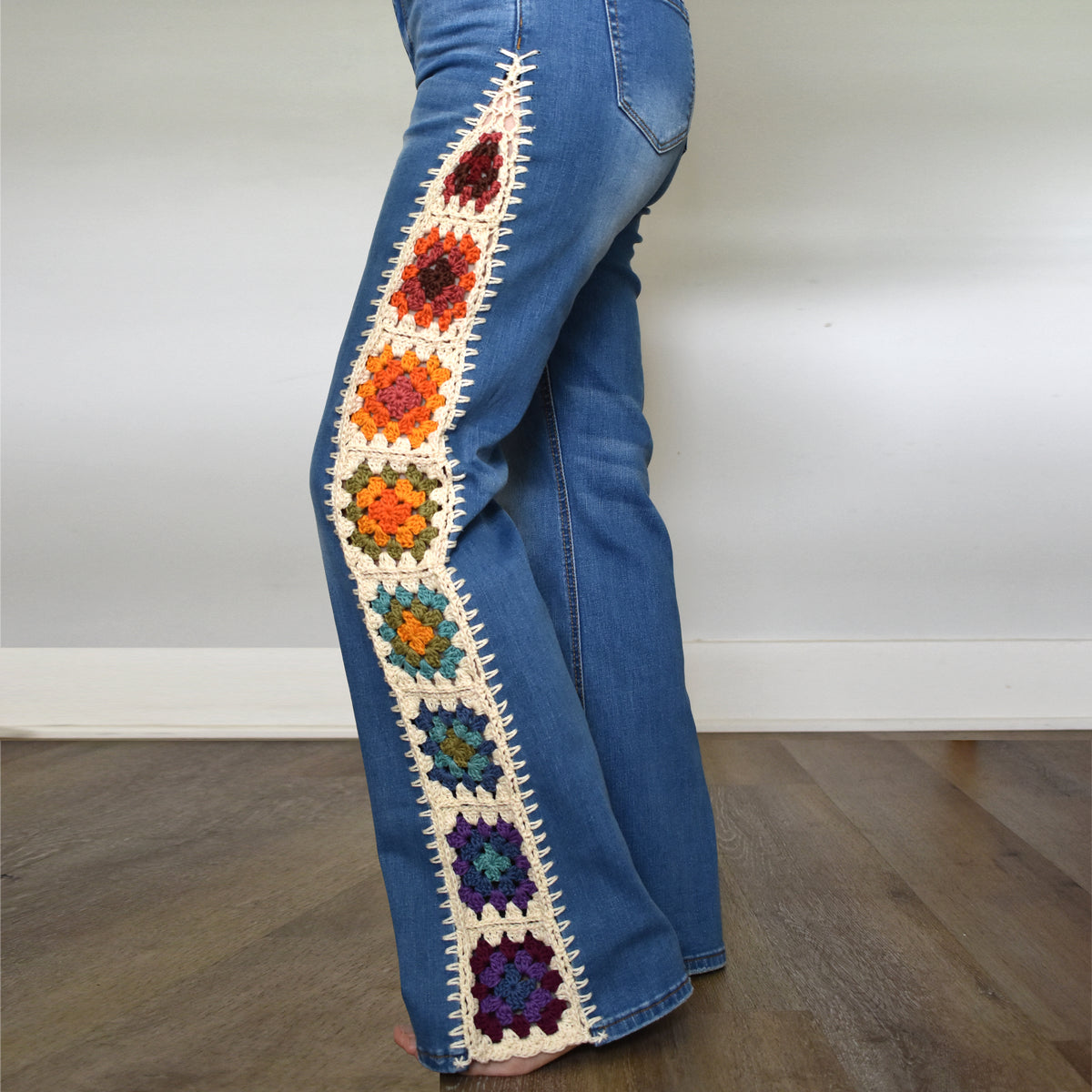 Blue denim jeans with a colorful crocheted floral panel running down the left leg, shown on a person standing on a wooden floor against a plain white wall.