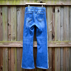 Blue denim jeans with white contrast stitching down the outer seams of the legs, shown from the back hanging on a wooden hanger against a weathered wooden fence.