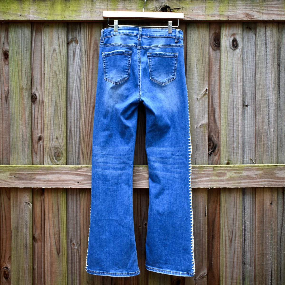 Blue denim jeans with white contrast stitching down the outer seams of the legs, shown from the back hanging on a wooden hanger against a weathered wooden fence.