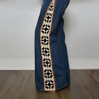 A close-up of a person's leg wearing blue denim jeans with a wide, decorative crochet stripe down the side in cream, black, and orange, standing on a wooden floor against a white wall.