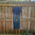 A pair of blue denim jeans with black and white geometric embroidered side panels, hanging from a wooden hanger against a wooden fence.