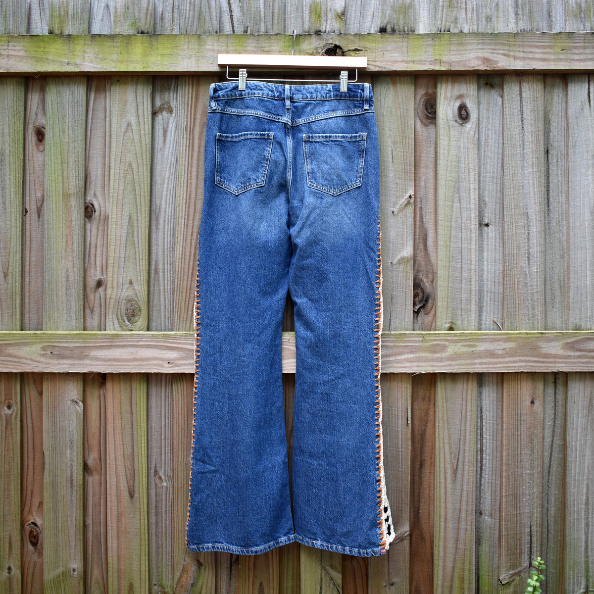 Blue denim jeans hanging on a wooden hanger against a weathered wooden fence, showing the back pockets and side stripe detailing.