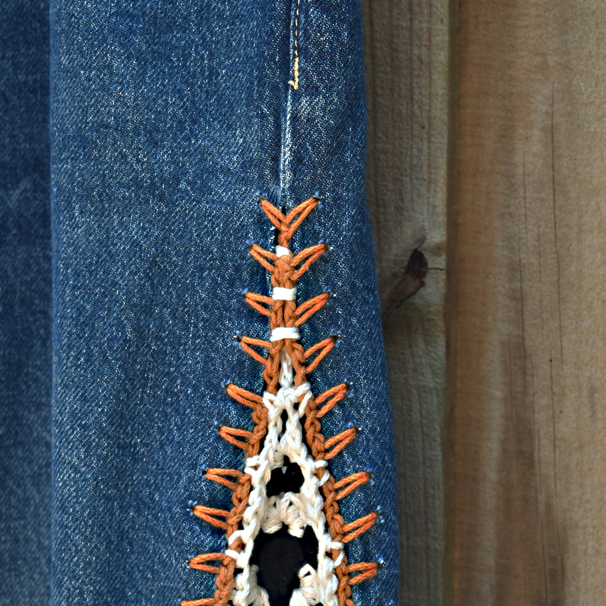 Close-up of a blue denim garment with a decorative orange and white crochet or knotted trim detail along a seam, set against a wooden background.