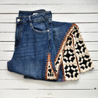 A pair of folded blue denim jeans with a black and cream crochet inserts and orange stitching, laid flat on a white wood plank background.