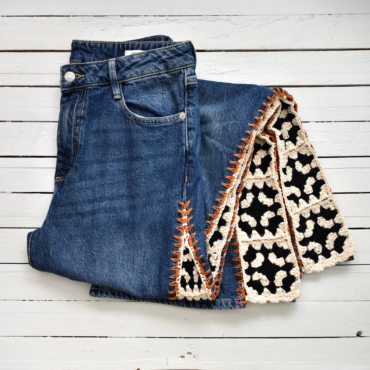 A pair of folded blue denim jeans with a black and cream crochet inserts and orange stitching, laid flat on a white wood plank background.
