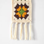 Crocheted wall hanging with colorful granny squares and tassels on a white background.