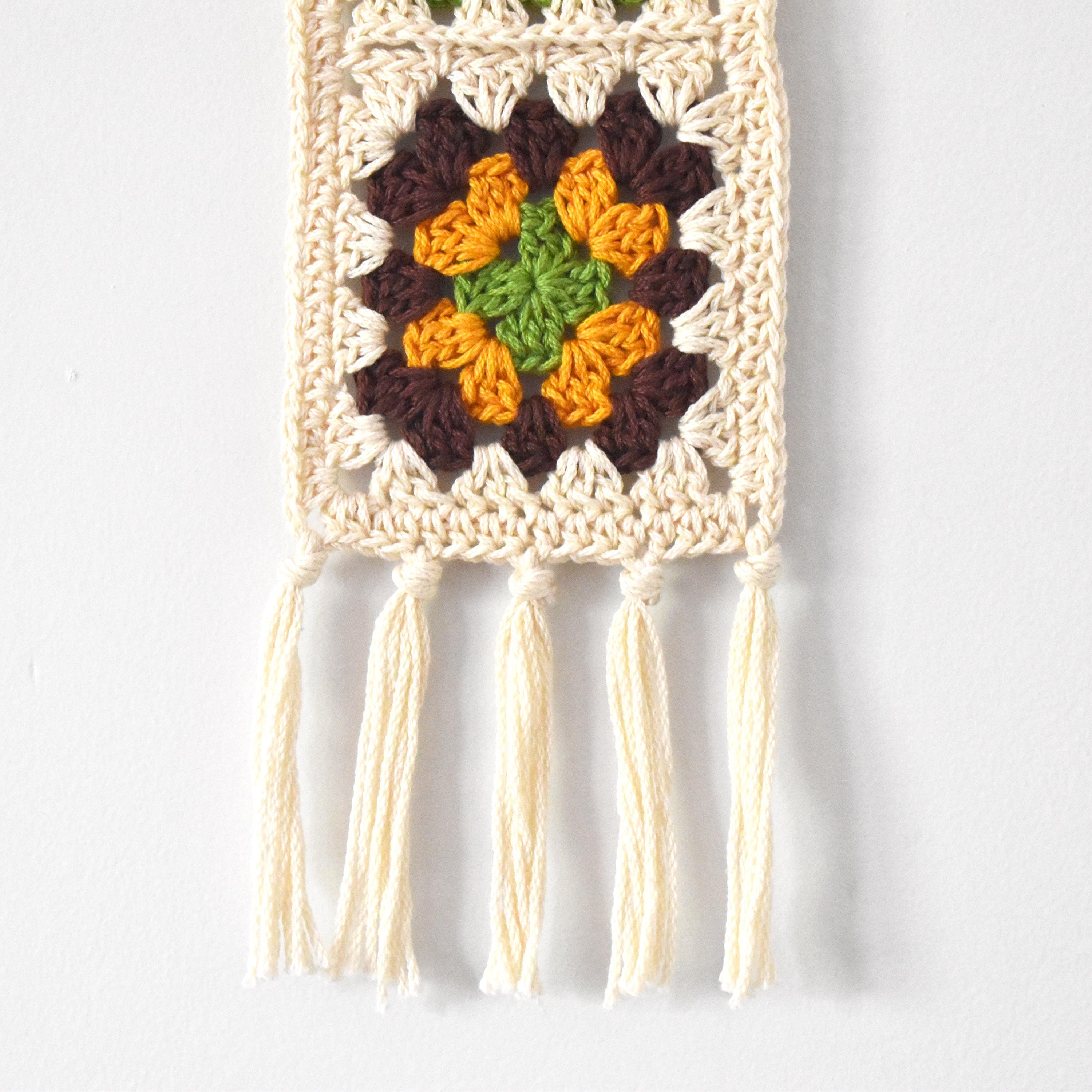 Crocheted wall hanging with colorful granny squares and tassels on a white background.