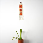 Crocheted wall hanging with geometric patterns next to a potted plant on a white background.