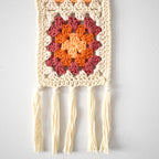 Crocheted wall hanging with a colorful granny square design and tassels on a white background.