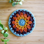 Colorful crocheted coaster on a wooden surface with green plants.