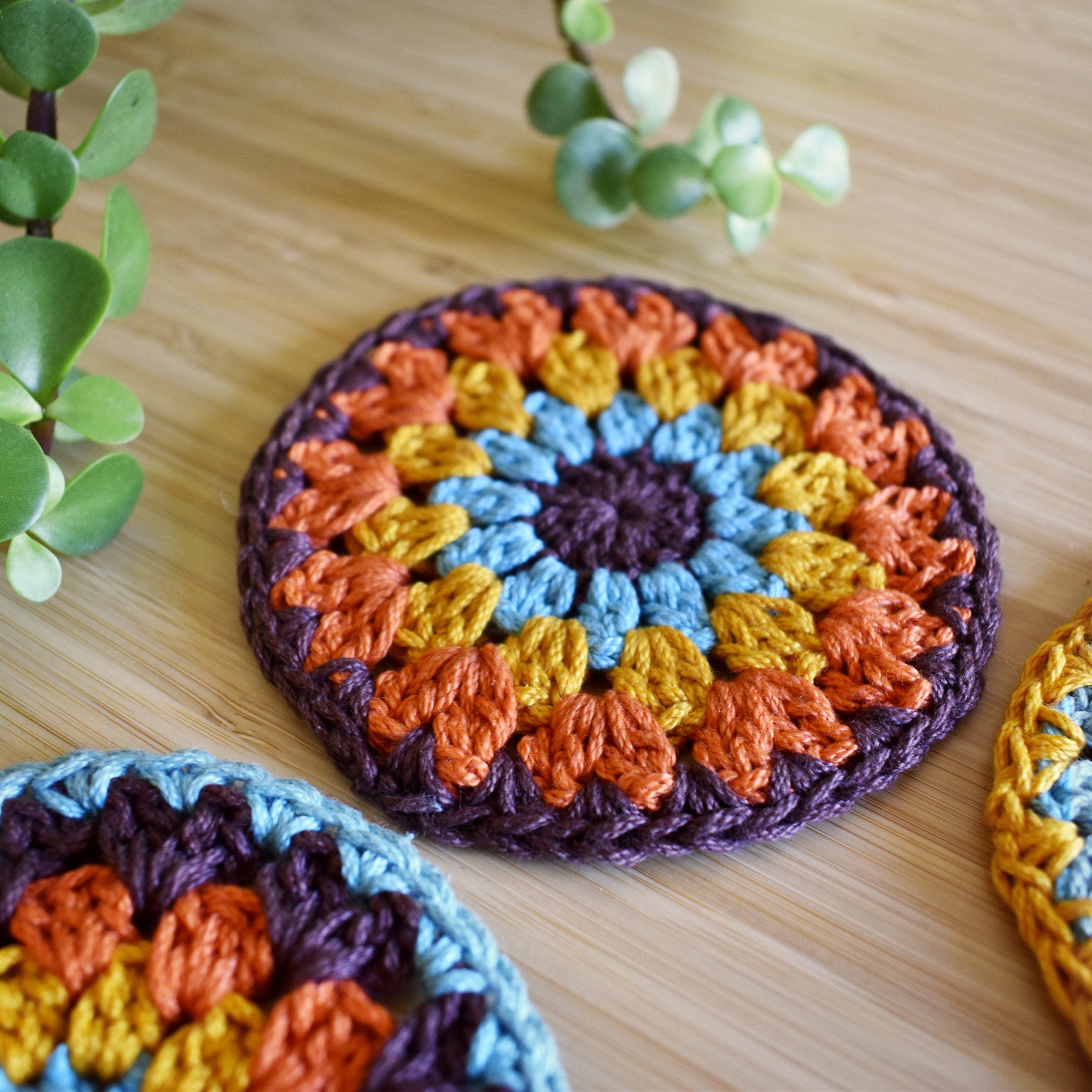 Colorful crocheted coasters on a wooden surface with green plants.