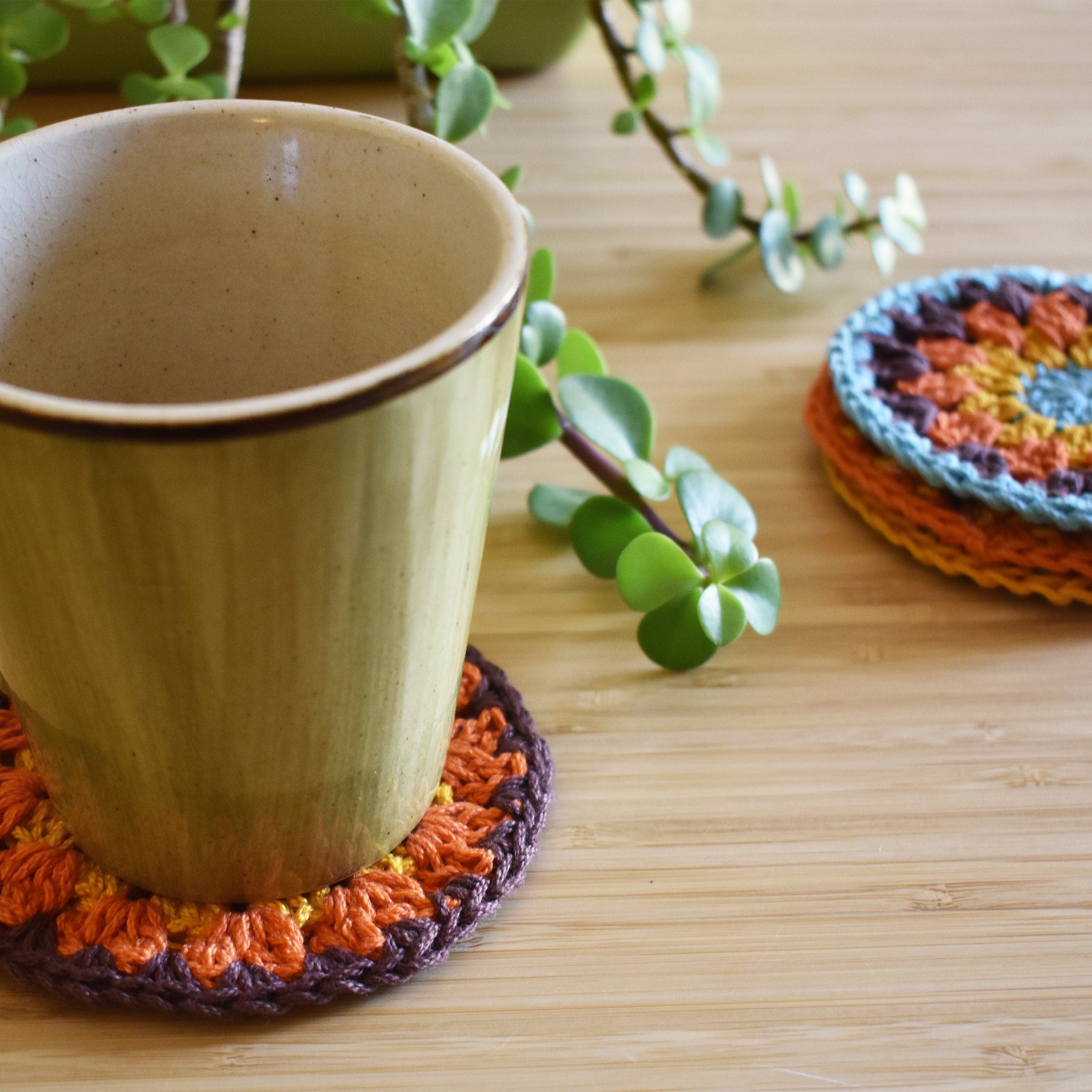 Ceramic cup with a colorful crocheted coaster on a wooden surface with a plant in the background.