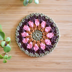Crocheted coaster with pink, brown, and green floral pattern on a wooden surface.