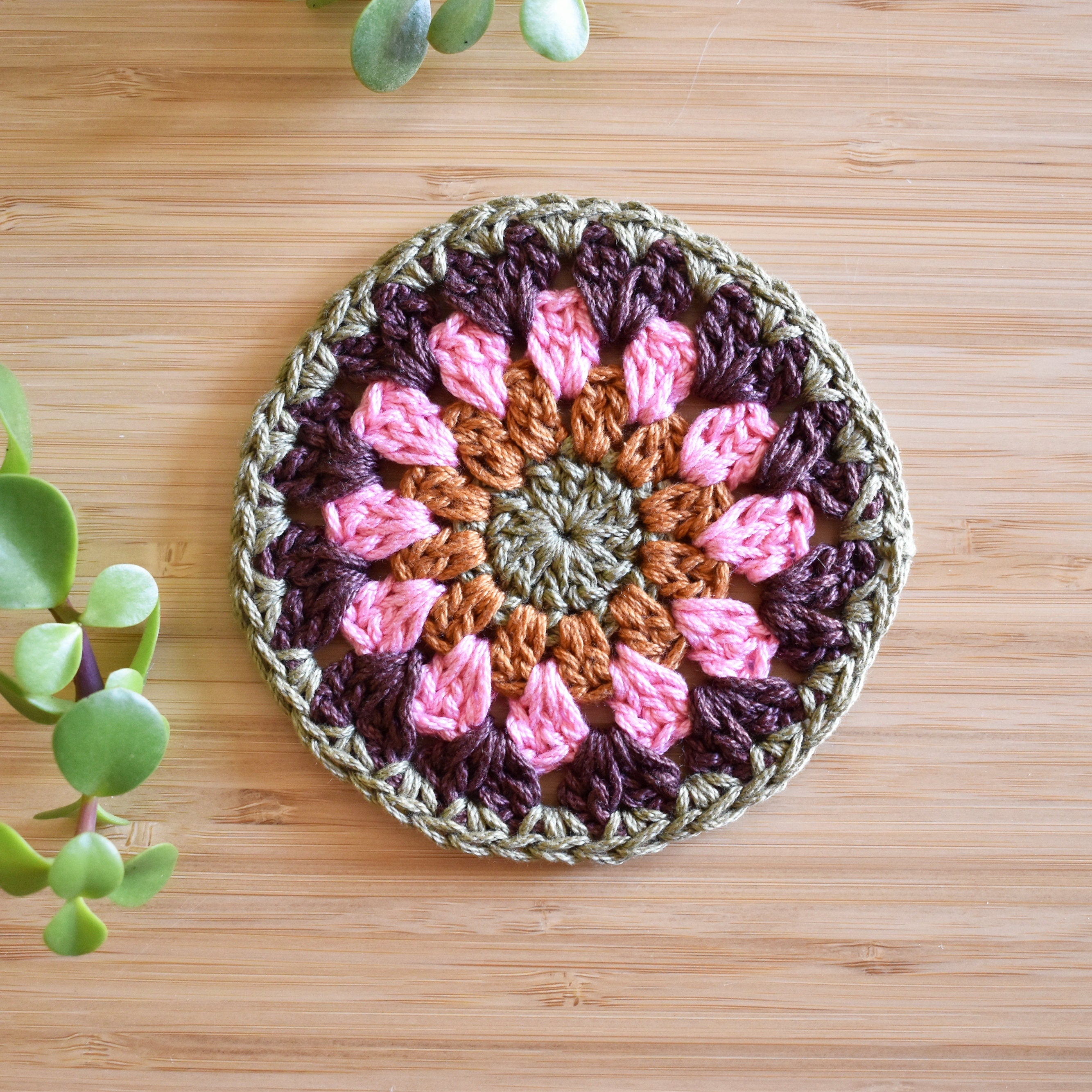 Crocheted coaster with pink, brown, and green floral pattern on a wooden surface.