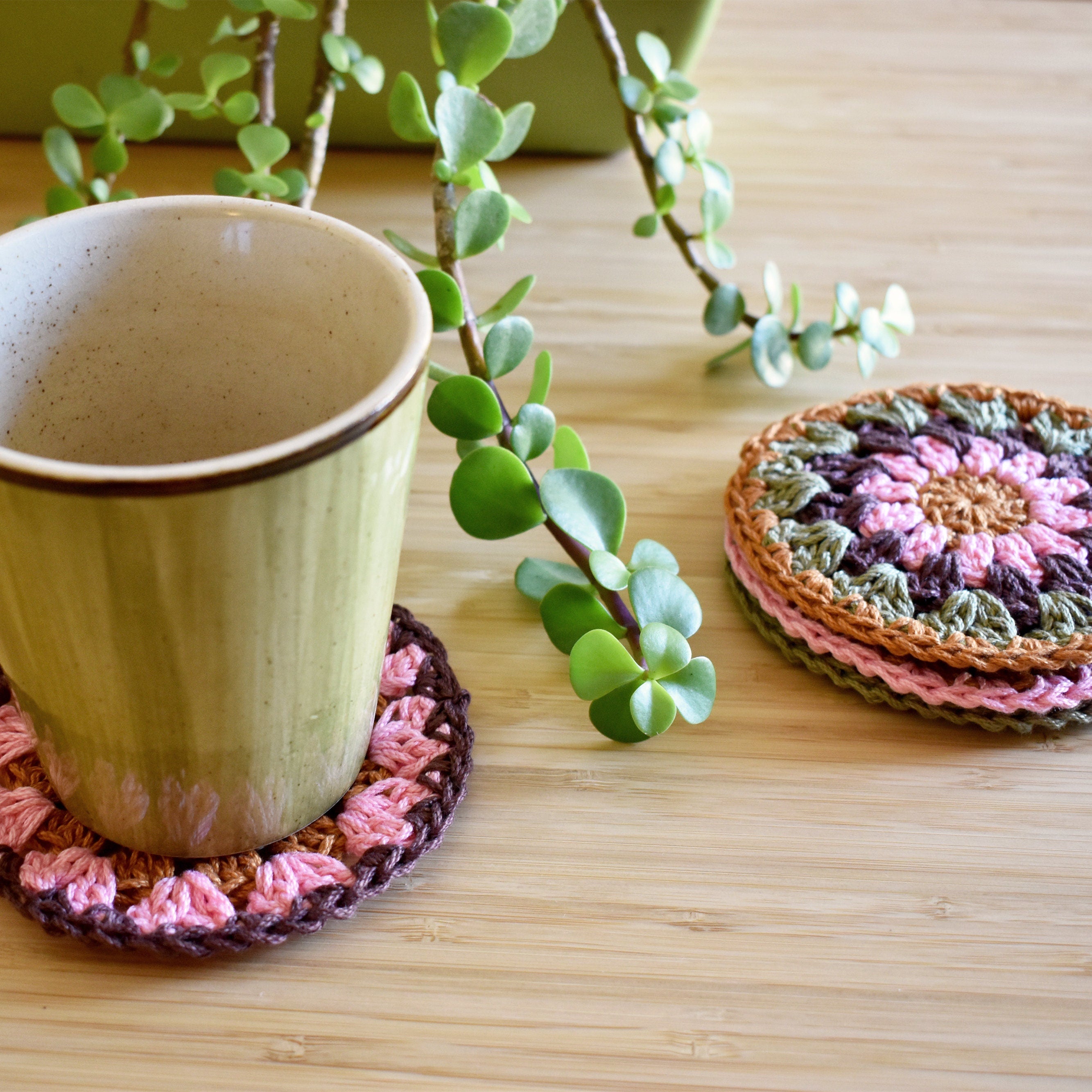 Ceramic cup with a colorful crocheted coaster on a wooden surface with a plant in the background.