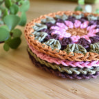 Stack of colorful crocheted coasters on a wooden surface with a plant in the background.