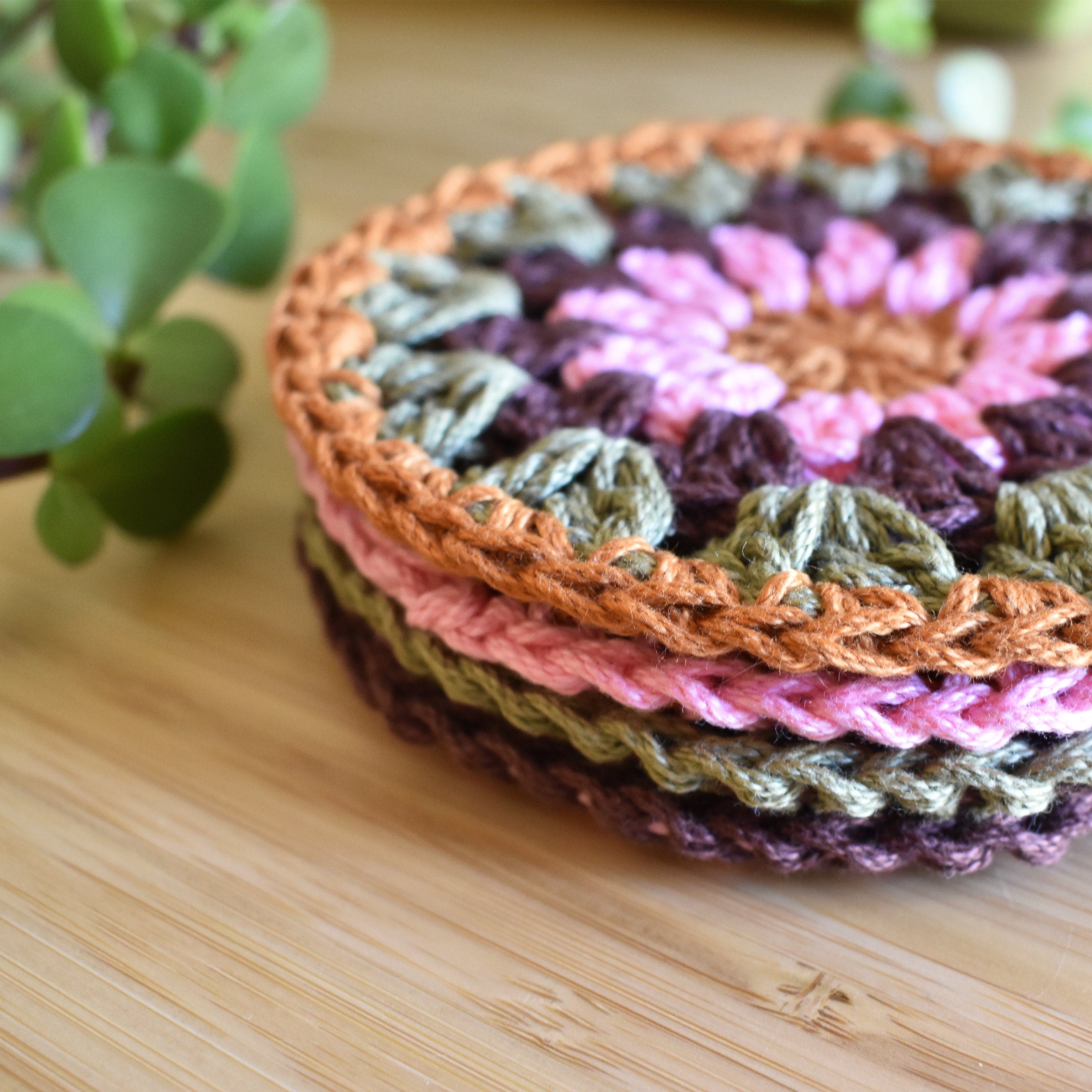 Stack of colorful crocheted coasters on a wooden surface with a plant in the background.