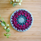 Crocheted coaster with purple and blue colors in a granny sunburst design on a wooden surface.