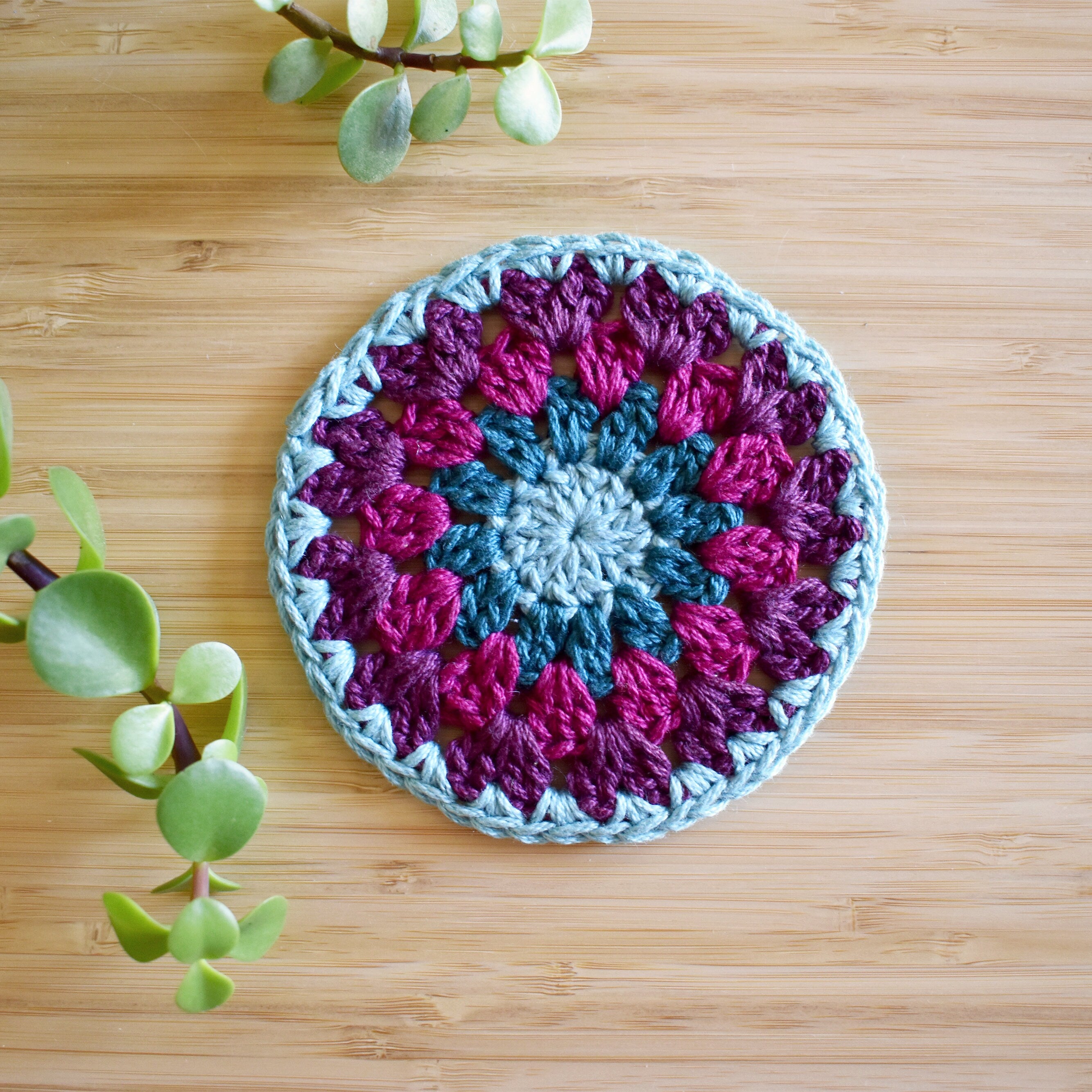 Crocheted coaster with purple and blue colors in a granny sunburst design on a wooden surface.