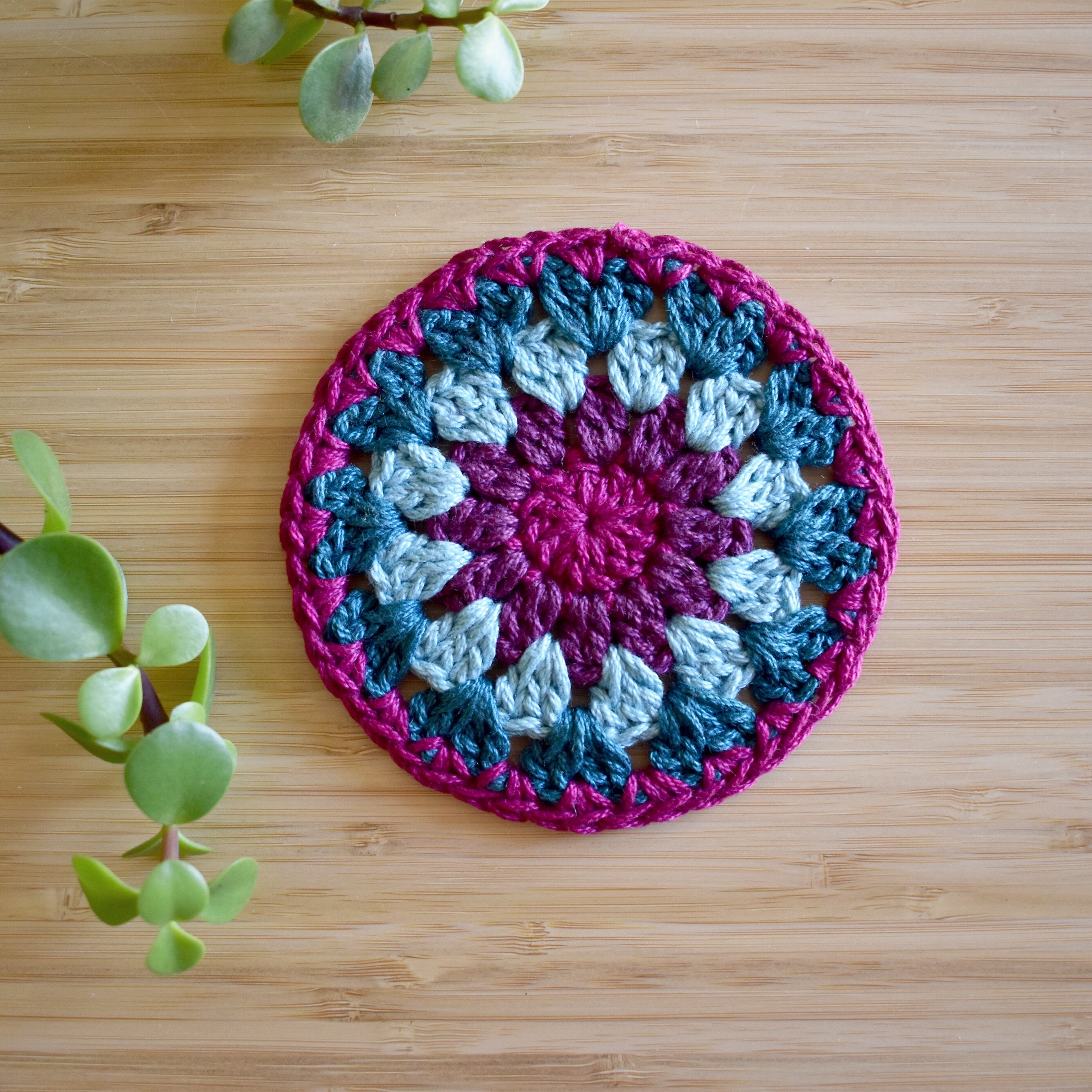 Round crochet coaster with purple and teal blue colors in a granny sunburst design on a wooden surface.