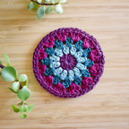 Purple and teal blue crocheted coaster with a colorful granny sunburst pattern on a wooden surface.