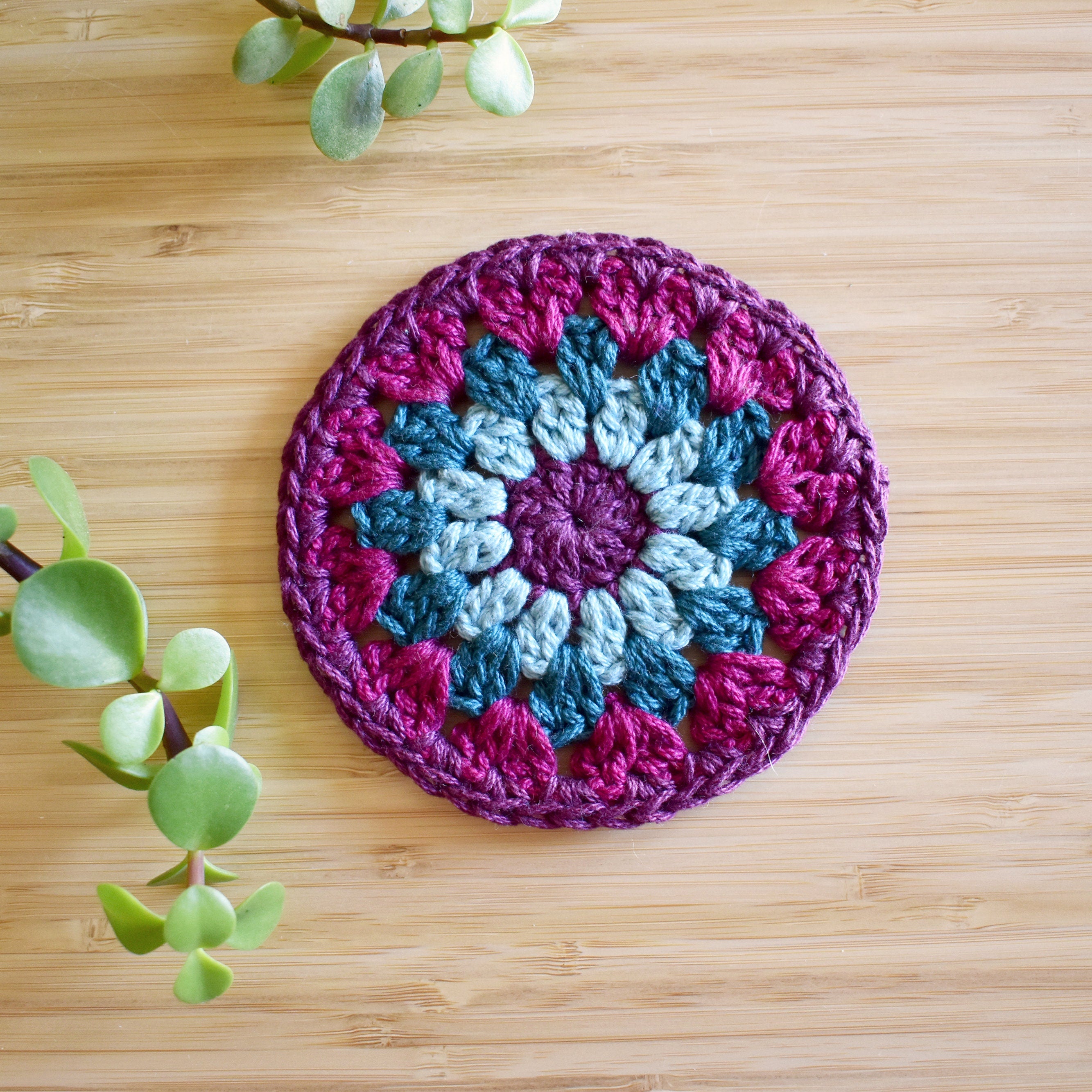 Purple and teal blue crocheted coaster with a colorful granny sunburst pattern on a wooden surface.
