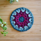Crocheted coaster with a purple and blue granny sunburst design on a wooden surface.