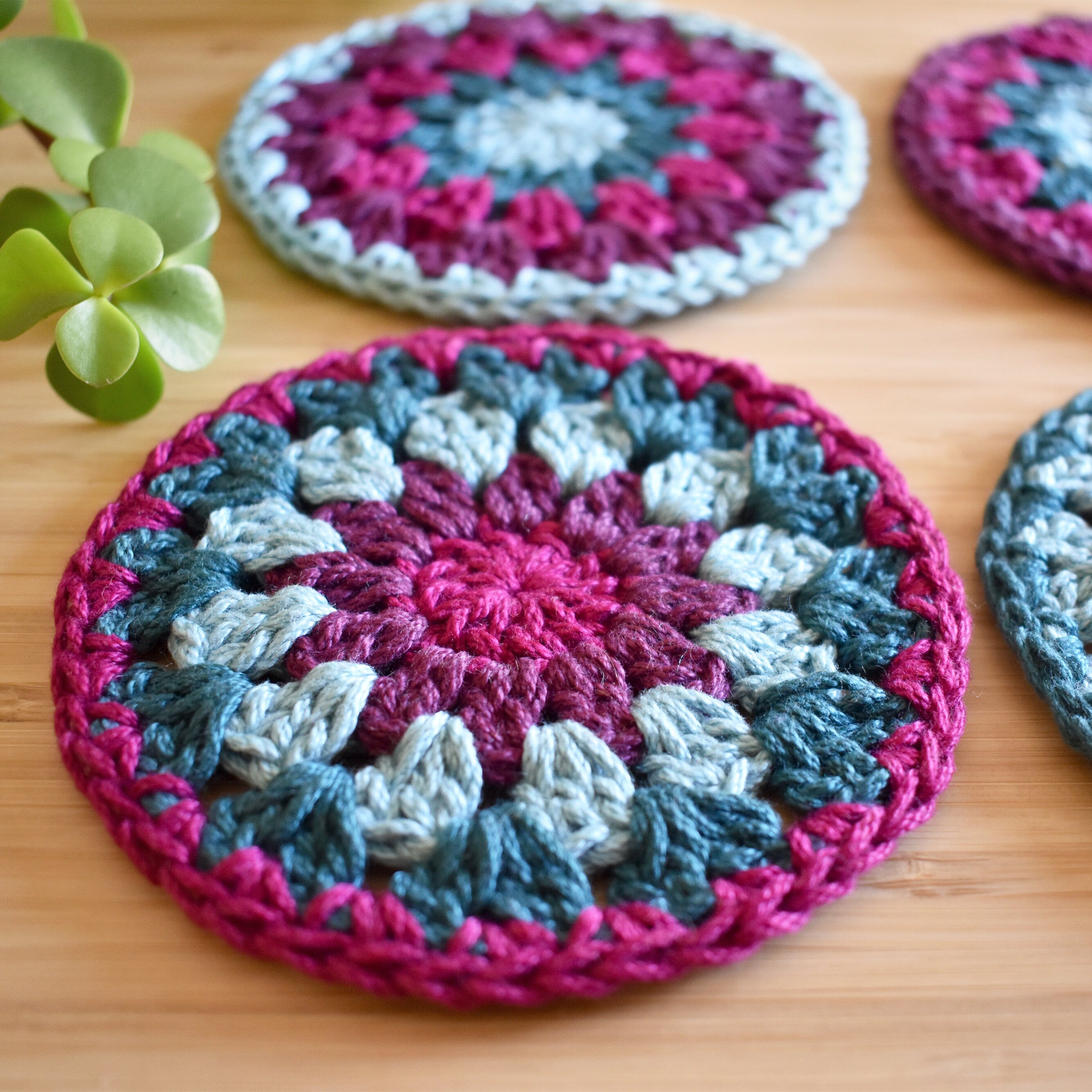Close-up view of colorful crocheted coasters on a wooden surface with a plant in the corner.
