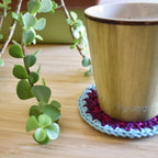 Ceramic mug on a colorful crocheted coaster with a plant in the background.