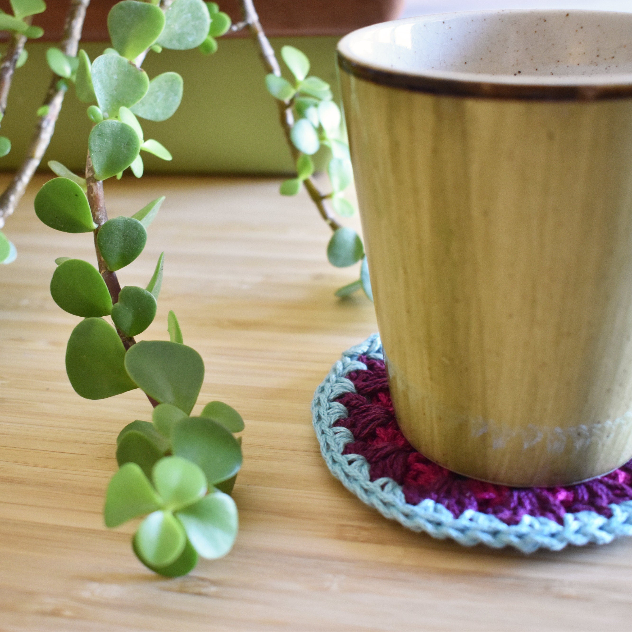 Ceramic mug on a colorful crocheted coaster with a plant in the background.