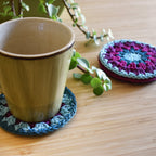 Ceramic mug with a crocheted coaster on a wooden surface, next to another coaster and a plant.